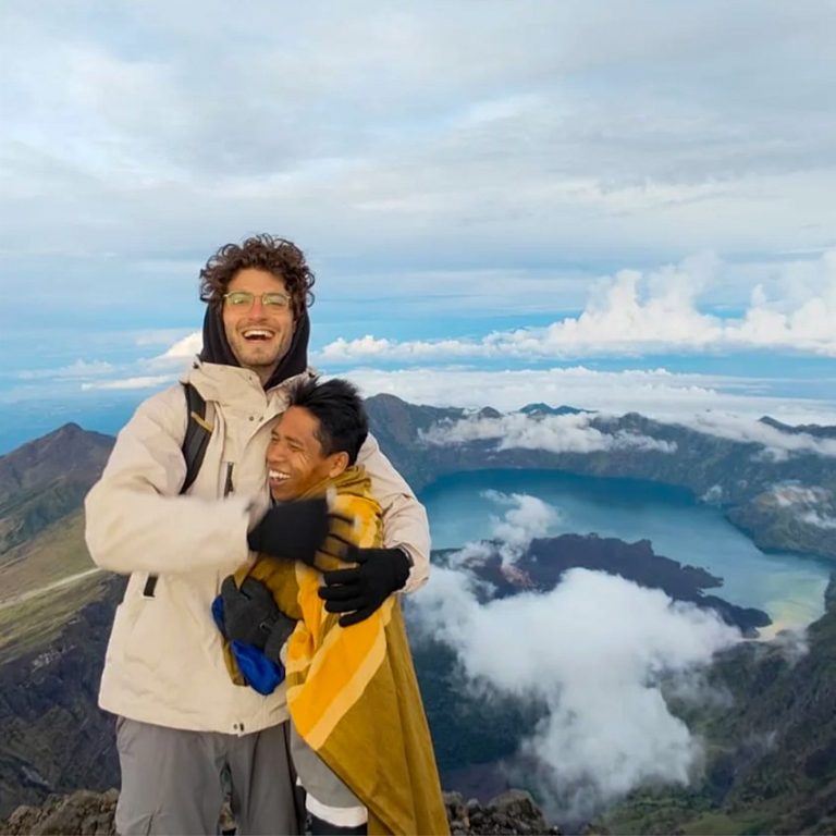 A happy trekker hugs his guide at the top of Mt. Rinjani, with the backdrop of the crater lake and daunting slopes, and rolling clouds.