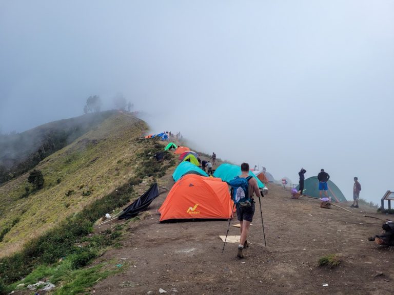 A group of hikers arrives at a mist-covered campsite along the Mt. Rinjani crater rim, where vibrant tents are set up on a narrow ridge with panoramic views obscured by dense fog, creating an atmospheric and rugged scene high above the clouds.