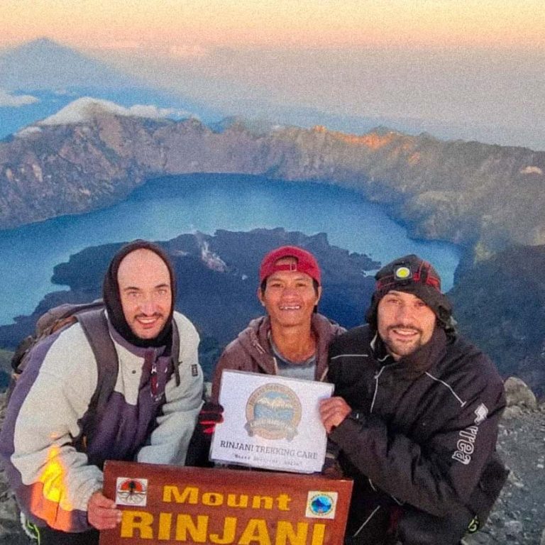 Two hikers pose with their guide and plaque at the summit of Mt. Rinjani, overlooking the crater lake.