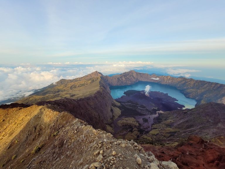 Stunning summit view from Mt. Rinjani showing the volcanic crater, Segara Anak lake, and a plume of smoke rising from the active volcano, surrounded by dramatic mountain ridges and a sea of clouds that emphasize the majesty of the Mt. Rinjani hike experience.