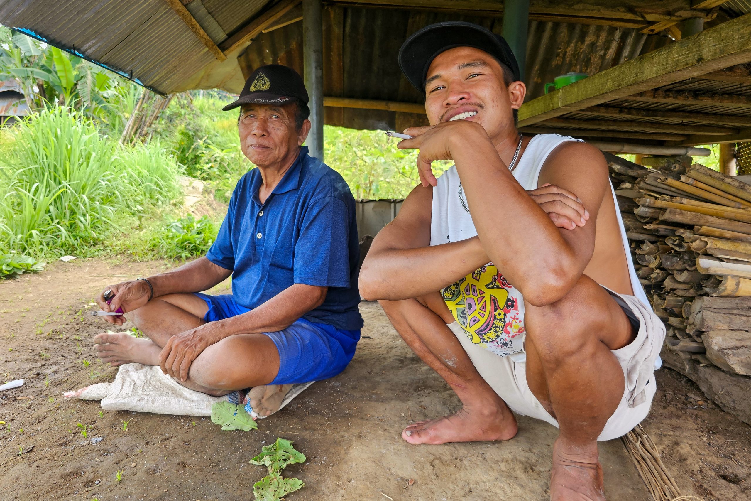 Two local men take a break under a simple shelter in a rural area near Mt. Rinjani, with one sitting cross-legged on the ground and the other crouching beside a small stack of firewood, both wearing casual clothes and hats while enjoying a relaxed conversation outdoors.