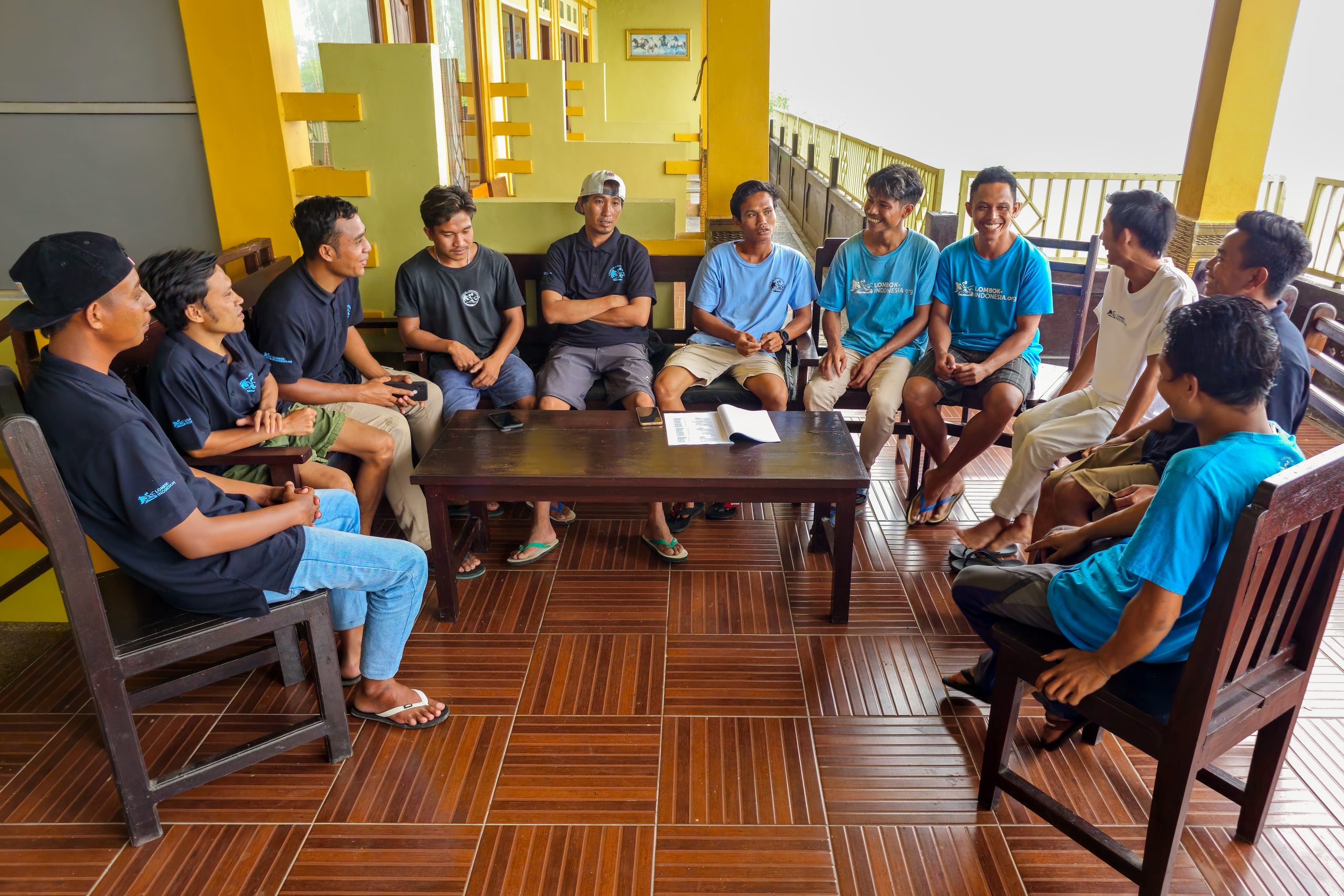 A group of local trekking staff members gather around a wooden table for a casual team meeting in a covered terrace space, with most of them wearing matching blue and black uniforms, smiling and engaged in friendly conversation as sunlight filters through the background.