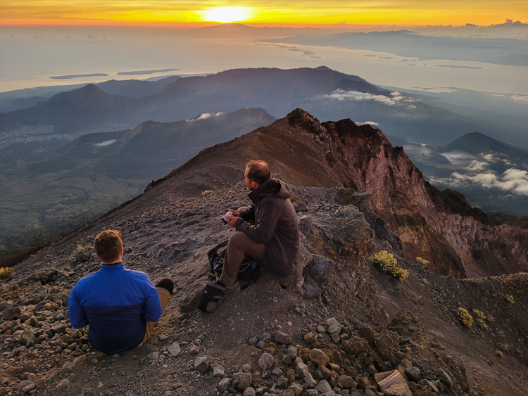 Two hikers resting at the summit of Mt. Rinjani during sunrise with vast coastal views in the distance, illustrating the reward and beauty awaiting those who complete the most challenging stretch of the Mt. Rinjani hike.