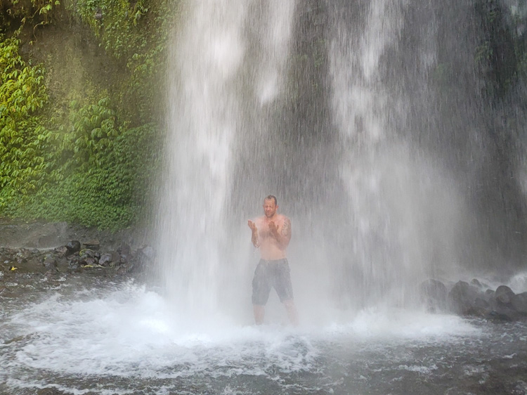 Man standing beneath a jungle waterfall surrounded by thick vegetation and mossy boulders, capturing a refreshing moment hikers often enjoy before or after the Mt. Rinjani hike as part of eco-tour experiences in Lombok.
