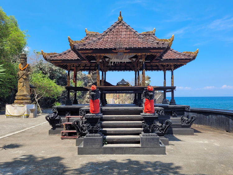 A traditional Balinese-style Hindu temple stands near the ocean with ornately carved black stone statues draped in red cloth, offering a peaceful spiritual stop for visitors who come to Lombok for cultural exploration as well as the popular Mt. Rinjani hike.