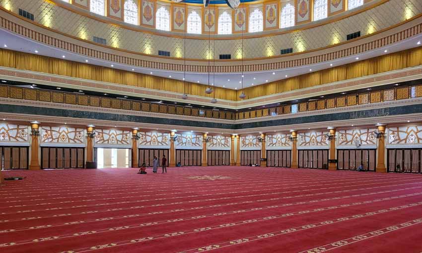 Interior view of the grand Islamic Center Mosque in Mataram, Lombok, showing its vast red-carpeted prayer hall, elegant chandeliers, and intricate architectural details, often visited as a cultural stop before or after a Mt. Rinjani hike through the island.