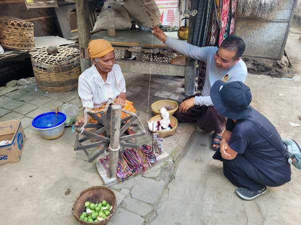 A local woman demonstrates traditional yarn spinning in a Sasak village while two visitors watch and learn, showcasing the cultural richness of Lombok and offering an authentic experience that often complements a Mt. Rinjani hike for travelers exploring the island.